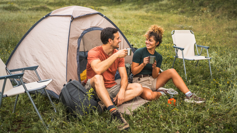 Two people sitting outside of a tent drinking from stainless steel mugs.
