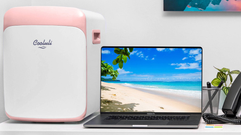 A pink and whilte Cooluli fridge on top of a desk, next to a laptop.