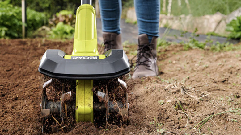 A person in jeans and brown boots using a Ryobi cultivator to till a garden