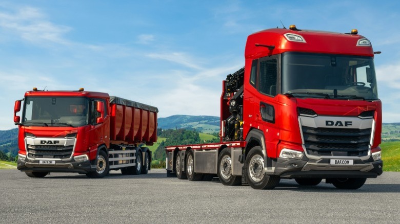 A red DAF semi-truck and vocational truck parked with green hills in the background