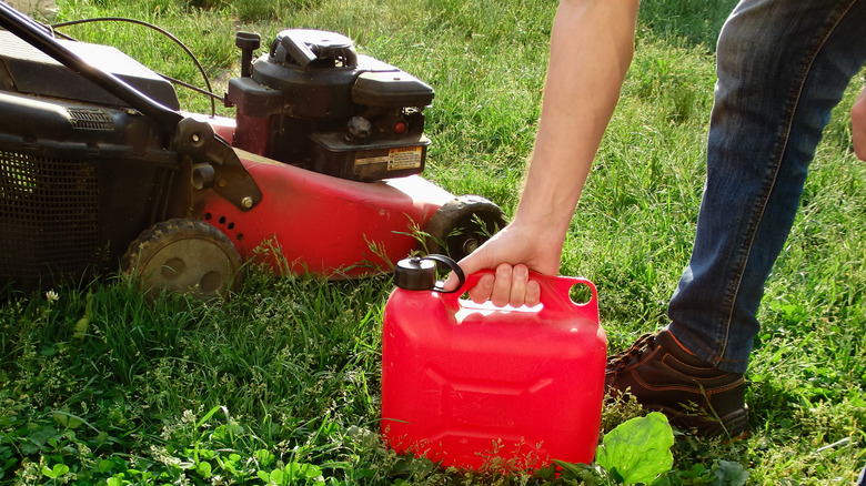 A person holding a gas canister beside a lawn mower.