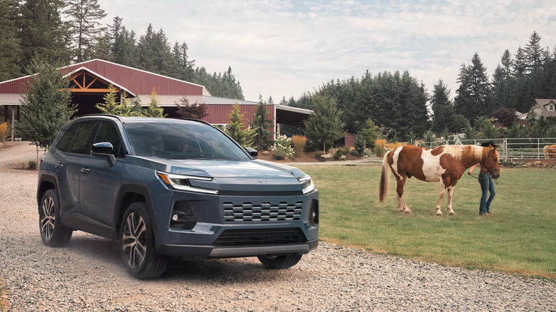 A blue-gray 2026 Toyota RAV4 crossover SUV standing on gravel outside a rural farm with a horse visible.