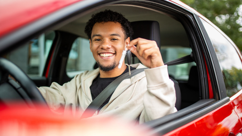 A man in a light colored hoodie behind the wheel of a red car, holding up the keys and smiling