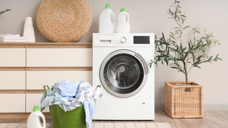 A modern laundry room with a white washing machine, green laundry basket filled with clothing, and white detergent bottles with green caps