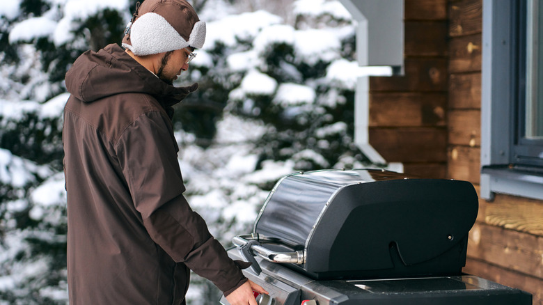 A man in a winter coat and hat with ear flaps turning on his grill outside near snow-covered trees