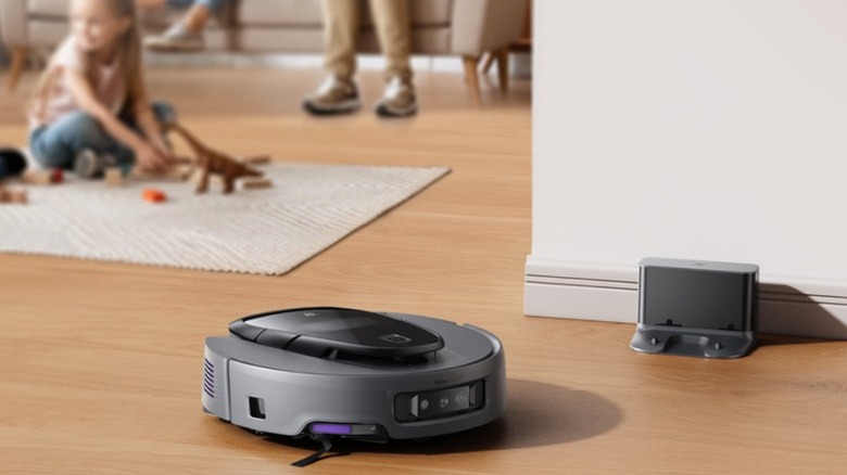 A gray 3i G10+ robot vacuum on wooden floors with child playing in the background