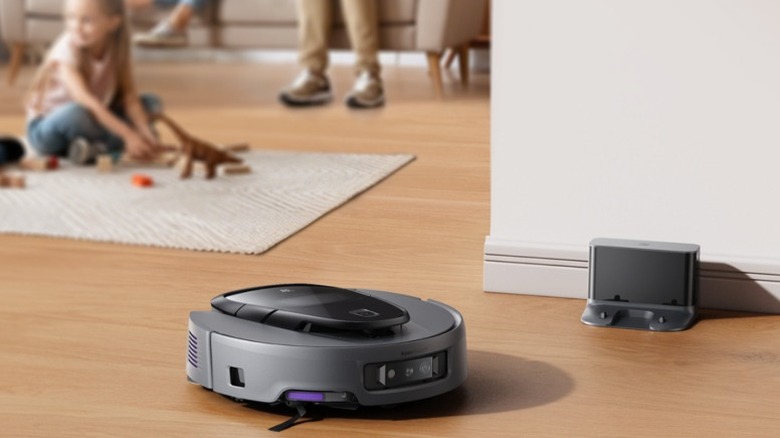 A gray 3i G10+ robot vacuum on wooden floors with child playing in the background