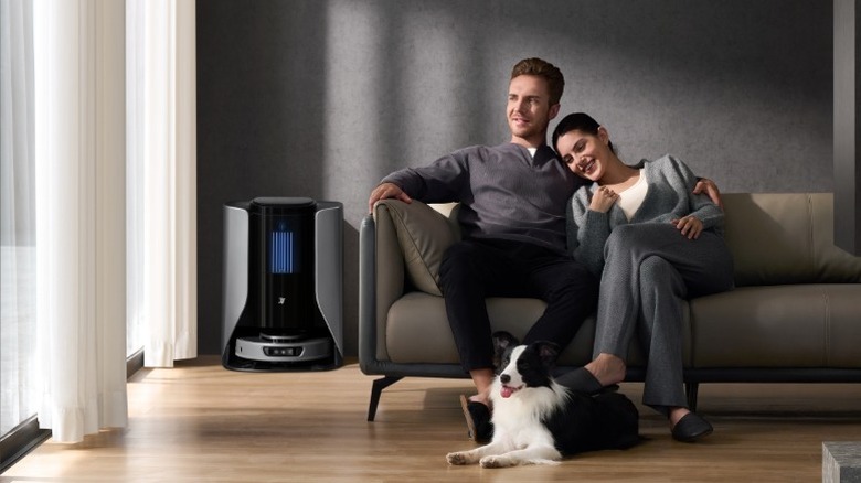 A woman and a man sitting on couch with dog with gray 3i S10 Ultra robot vacuum in the corner