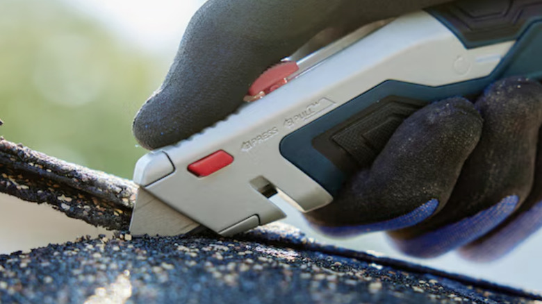 Close-up of a gloved hand using a Bosch utility knife to cut a roof shingle