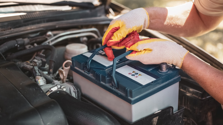 A person wearing gloves is handling a car battery inside an engine bay, with the sun in the background.