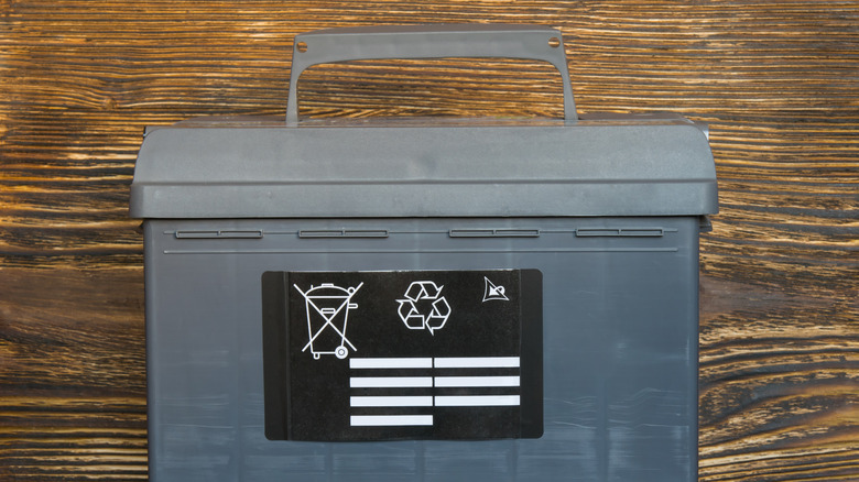 A gray car battery pictured in front of a wooden background, with a black sticker on it.
