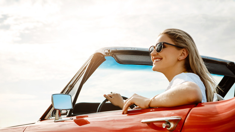 A smiling blond woman wearing sunglasses and driving a vintage red convertible
