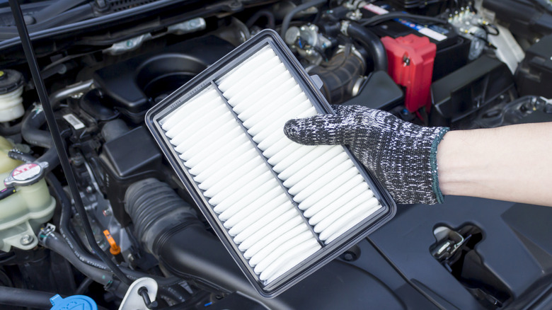 A gloved hand holding a clean air filter over a car's open engine compartment