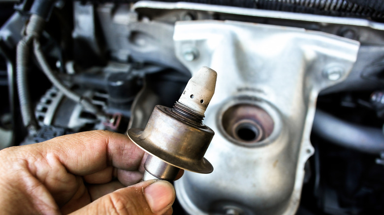 A man's hand holding an oxygen sensor over an open engine compartment