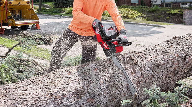 A worker using a Milwaukee M18 Fuel 20-inch dual-battery chainsaw on a fallen tree in a residential neighborhood