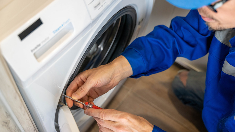 A man in a blue jacket and baseball cap using a screwdriver to repair a door on a washing machine