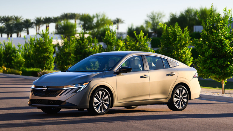 A gold 2027 Nissan Sentra parked on pavement with rows of trees in the background