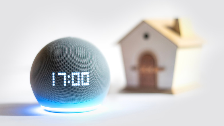 An Amazon Echo Dot displaying the time placed next to a model house