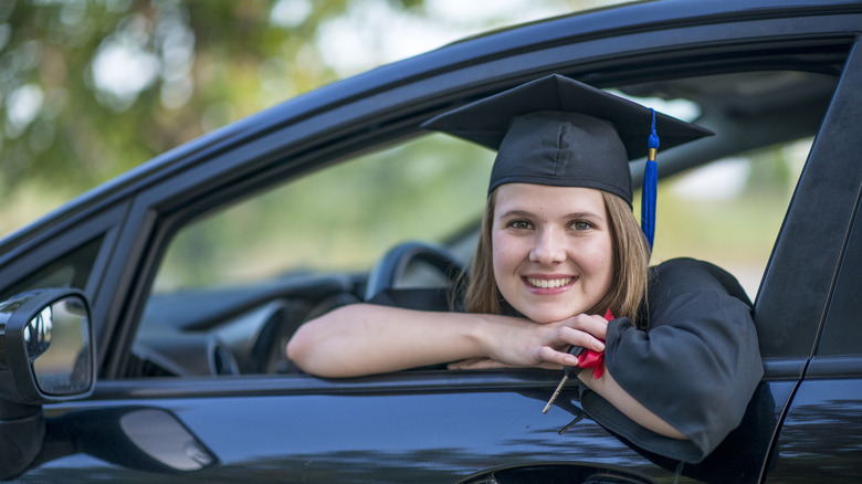 A smiling woman in a black graduation cap and gown in the driver's seat of a black vehicle