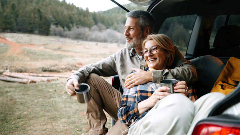 A smiling couple sitting in the open rear of their SUV with camping mugs in a remote site