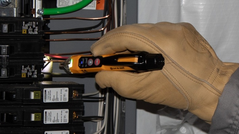 A person wearing work gloves using the Klein Tools non-contact voltage tester on a circuit breaker panel.