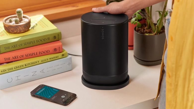 A person touching a Sonos Move 2 portable speaker surrounded by books and a smartphone.
