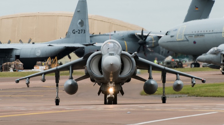 gray-colored AV-8B Harrier sitting on a runway with other military planes in the background