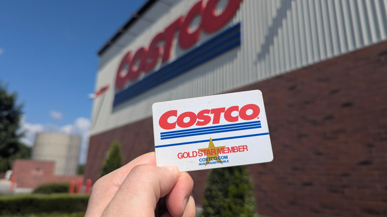 A close-up of a hand holding a Costco Gold Star Member membership card in front of a Costco store