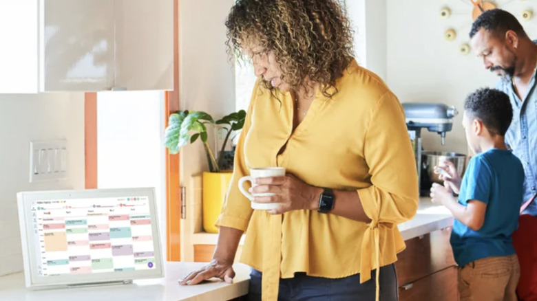 A woman in yellow holding a coffee mug looks at her Skylight calendar in her kitchen with her husband and son standing behind her