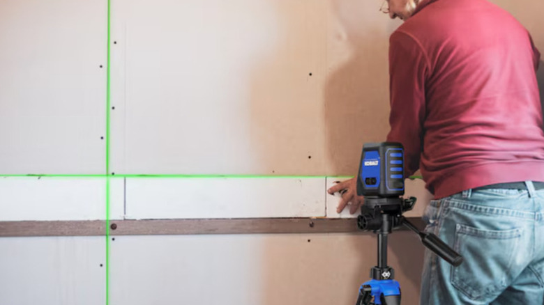 Kobalt self-leveling cross-line laser level mounted on tripod with green laser guiding man hanging drywall