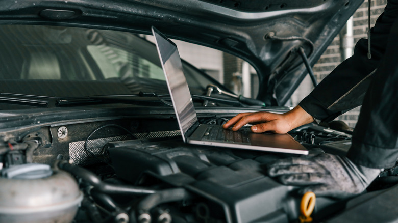 A mechanic using a laptop under the hood of an unidentified vehicle, presumably to diagnose some type of fault
