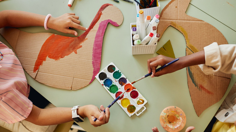Two children working on craft projects with cardboard shapes and paint at a green table