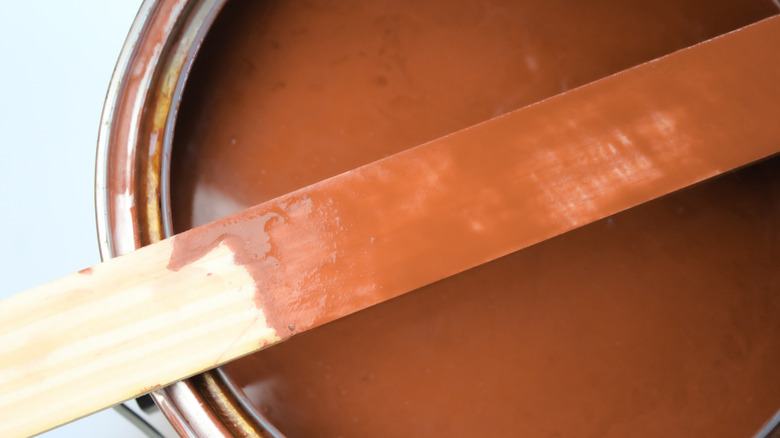 Close-up of an open can of brown paint with a paint-covered stirring stick laying across the top