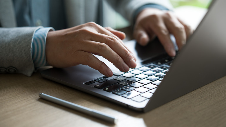 A person typing on a laptop that's placed on a wooden desk with a pen on the side