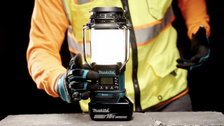 A man in a yellow safety vest, orange shirt and blue gloves placing a lit Makita brand lantern on a stone surface