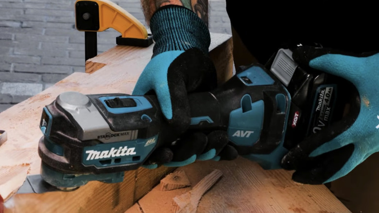 A man wearing blue gloves using a Makita Oscillating Multi‑Tool on a wood surface