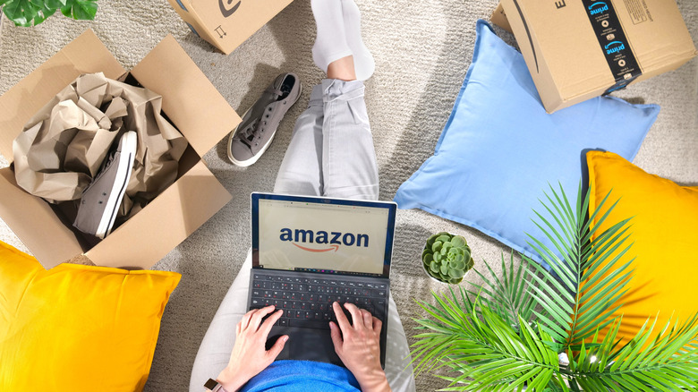 A person sitting on a carpeted floor using a laptop to shop on Amazon while surrounded by Amazon boxes and colorful pillows