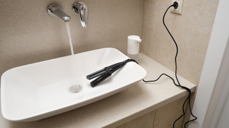 A hair tool plugged in and sitting on the edge of a sink with running water in a bathroom