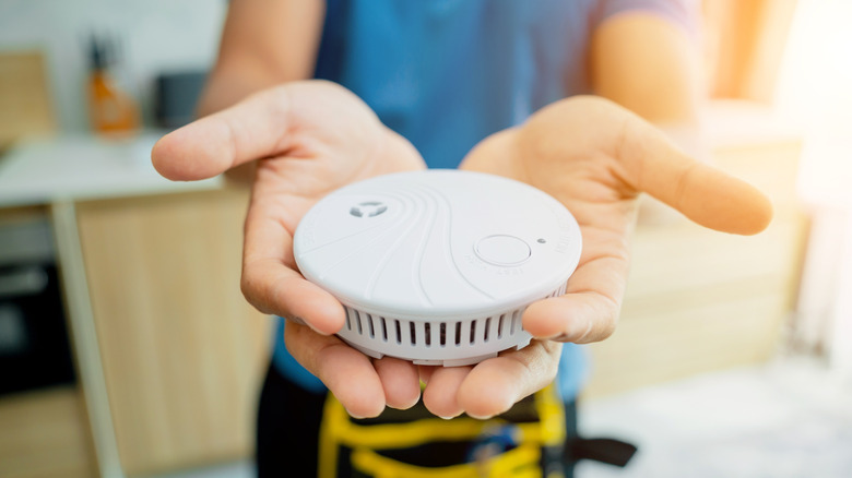 A person in a blue shirt wearing a tool belt holding out a white smoke detector in two hands prior to installation