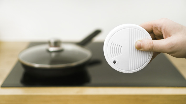 A hand holding a white smoke detector in front of a cooktop with an empty lidded pan sitting on it