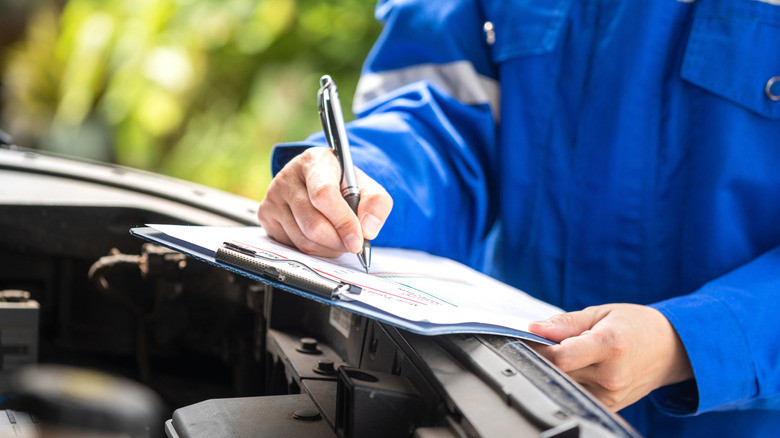 A man in blue coveralls writing on a clipboard propped up on the open hood of a vehicle