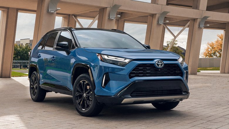 A blue 2025 Toyota RAV4 Hybrid, front 3/4 view, modern structure in the background