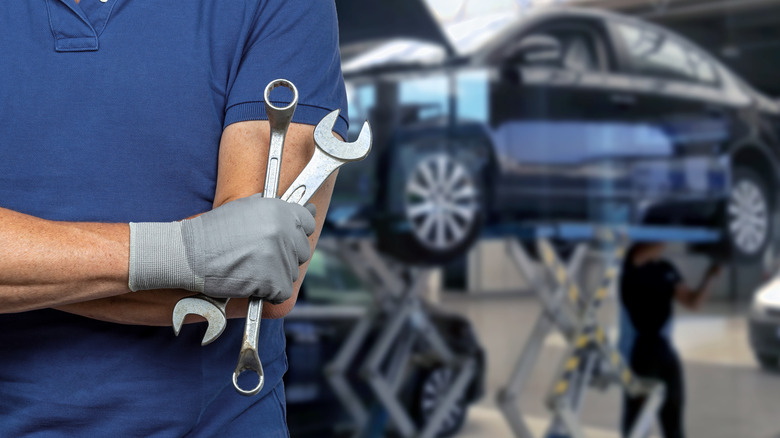 A person in a blue shirt holding two wrenches in an auto shop, car on a lift in the background