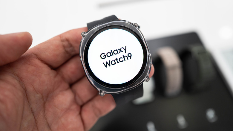 A hand holds a Samsung Galaxy Watch 9 that's displaying its name on a white screen.