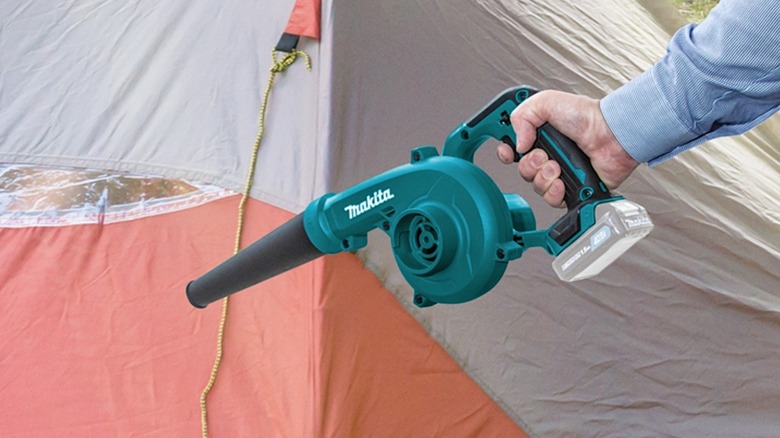 A hand holding a Makita 12V blower in front of a camping tent.