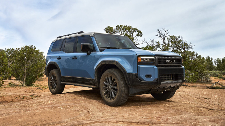 A light blue 2024 Toyota Land Cruiser parked in a rocky environment with trees in the background.