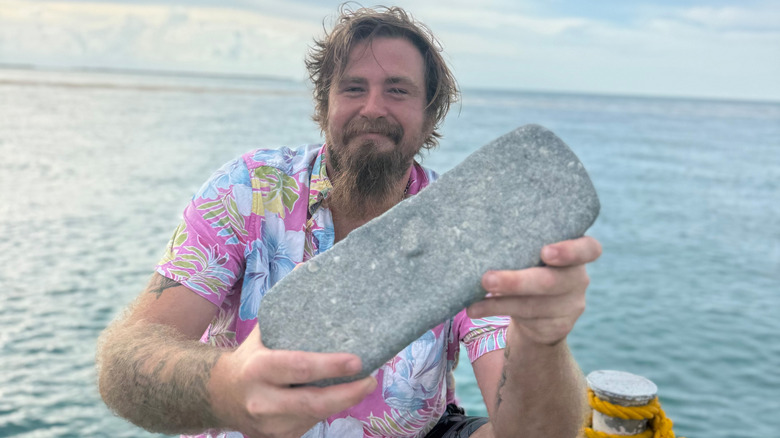 Bearded man with a pink Hawaiian shirt holding a large silver bar on a boat at sea.