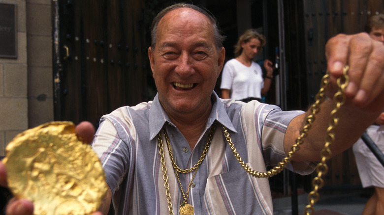 Older man holding a gold chain in one hand and large round gold coin shaped object in the other.