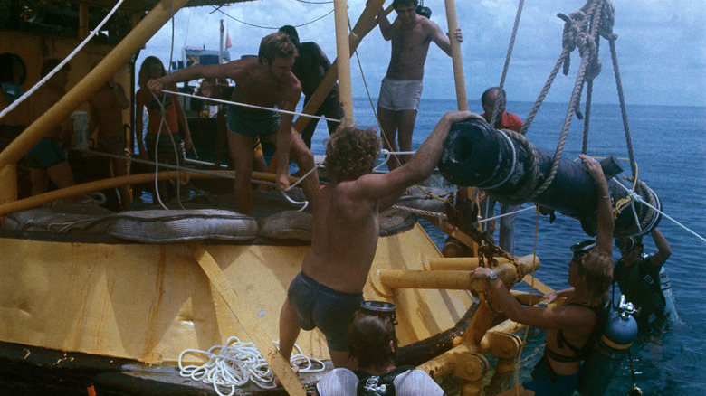 Multiple people pulling an old cannon out of the water in the ocean using a pulley.
