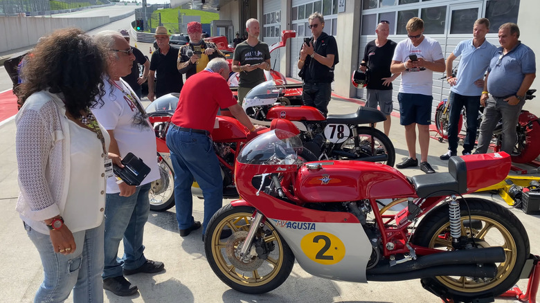 A crowd of onlookers admiring a red and white MV Agusta 500 GP motorbike in the pit lane of a race track.